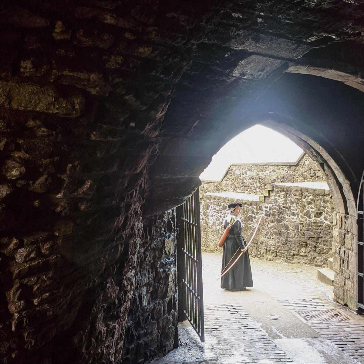 A woman dressed in a black 16th century kirtle, framed by a stone archway with wrought iron gates; she is holding a longbow. The overall effect is vaguely menacing, like she might decide to shoot an arrow in someone's face.